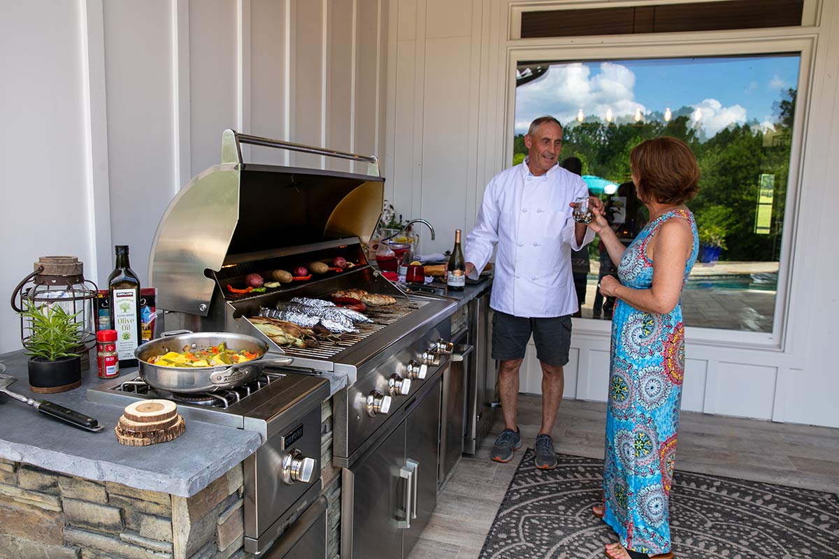 a couple standing by their linear outdoor kitchen cooking food on grill